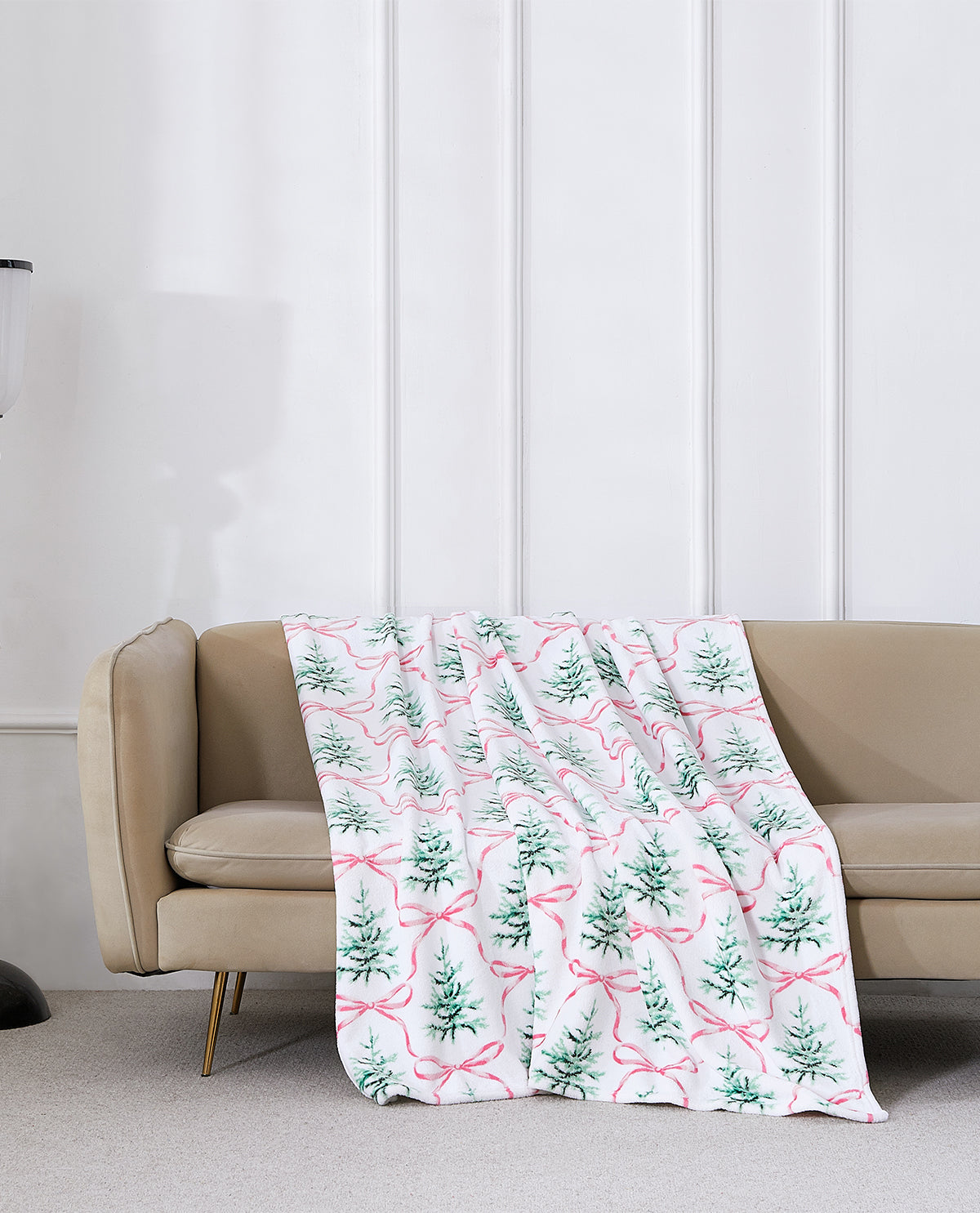 A white throw blanket draped over a beige sofa, featuring a repeating pattern of green pine trees and pink bows, set against a white paneled wall.