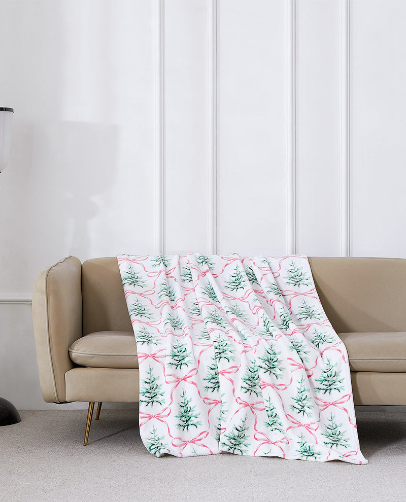 A white throw blanket draped over a beige sofa, featuring a repeating pattern of green pine trees and pink bows, set against a white paneled wall.