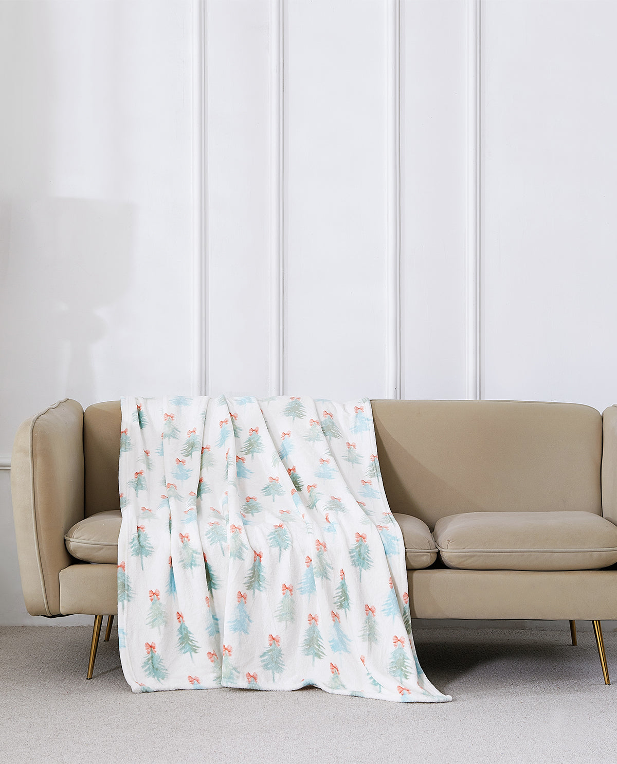 A white throw blanket with a light blue and coral Christmas tree pattern is draped over a beige sofa in a modern living room setting.