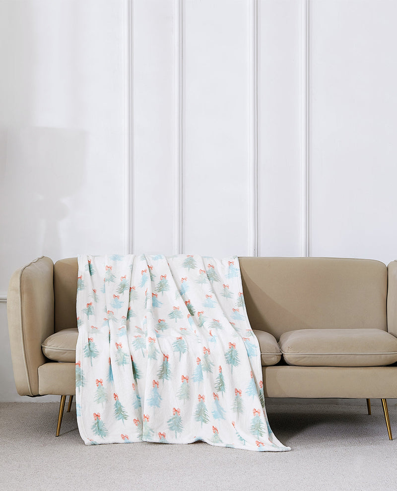 A white throw blanket with a light blue and coral Christmas tree pattern is draped over a beige sofa in a modern living room setting.
