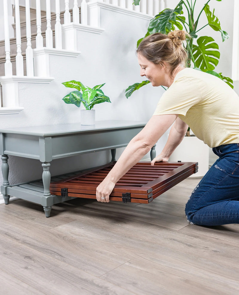 A woman kneels beside a gray console table in a bright indoor room, pulling out a dark wood slatted storage tray from the table's lower shelf.