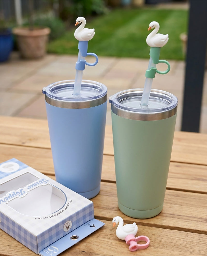 Two insulated travel tumblers with stainless steel lids and swan-shaped straws, shown on a wooden table outdoors. One tumbler is light blue and the other is pale green.