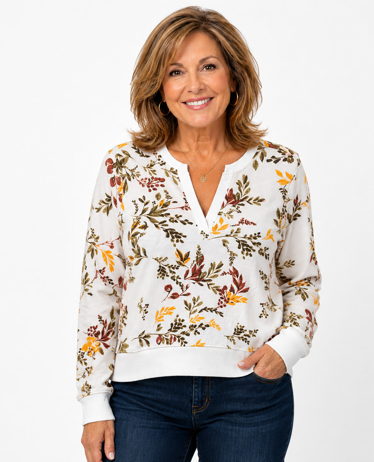 A smiling woman wearing a white long-sleeve knit top with a dense autumn floral and leaf print in brown, rust, and mustard yellow, paired with dark blue jeans, against a plain white studio background.