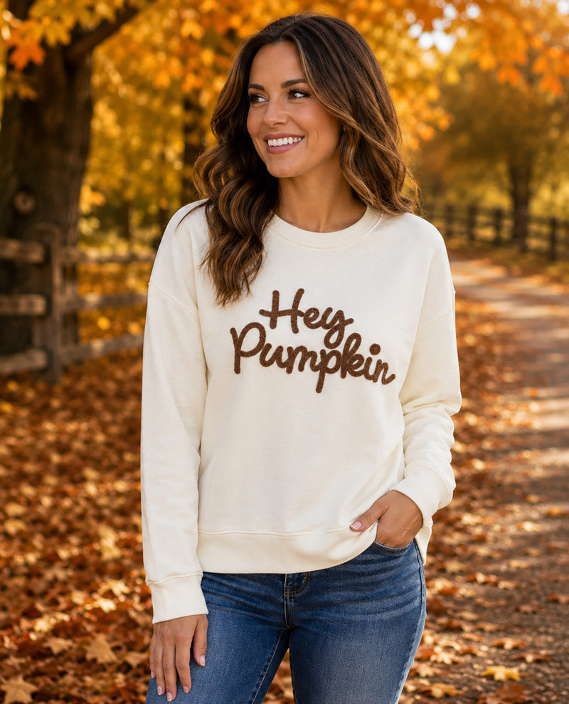 A woman wearing a cream-colored crewneck sweatshirt with 'Hey Pumpkin' written in brown script across the chest, standing outdoors on a leaf-covered path during autumn.