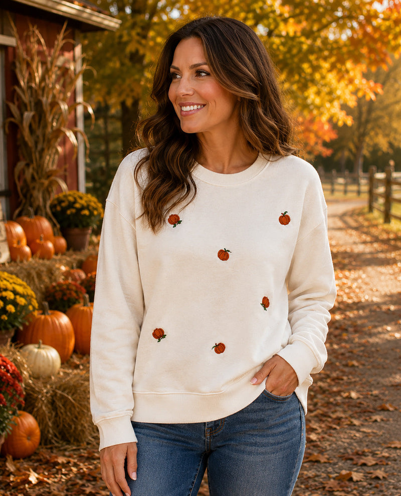 A woman wearing a cream-colored crewneck sweatshirt with embroidered orange pumpkins, standing in an autumn outdoor setting with pumpkins and fall foliage.