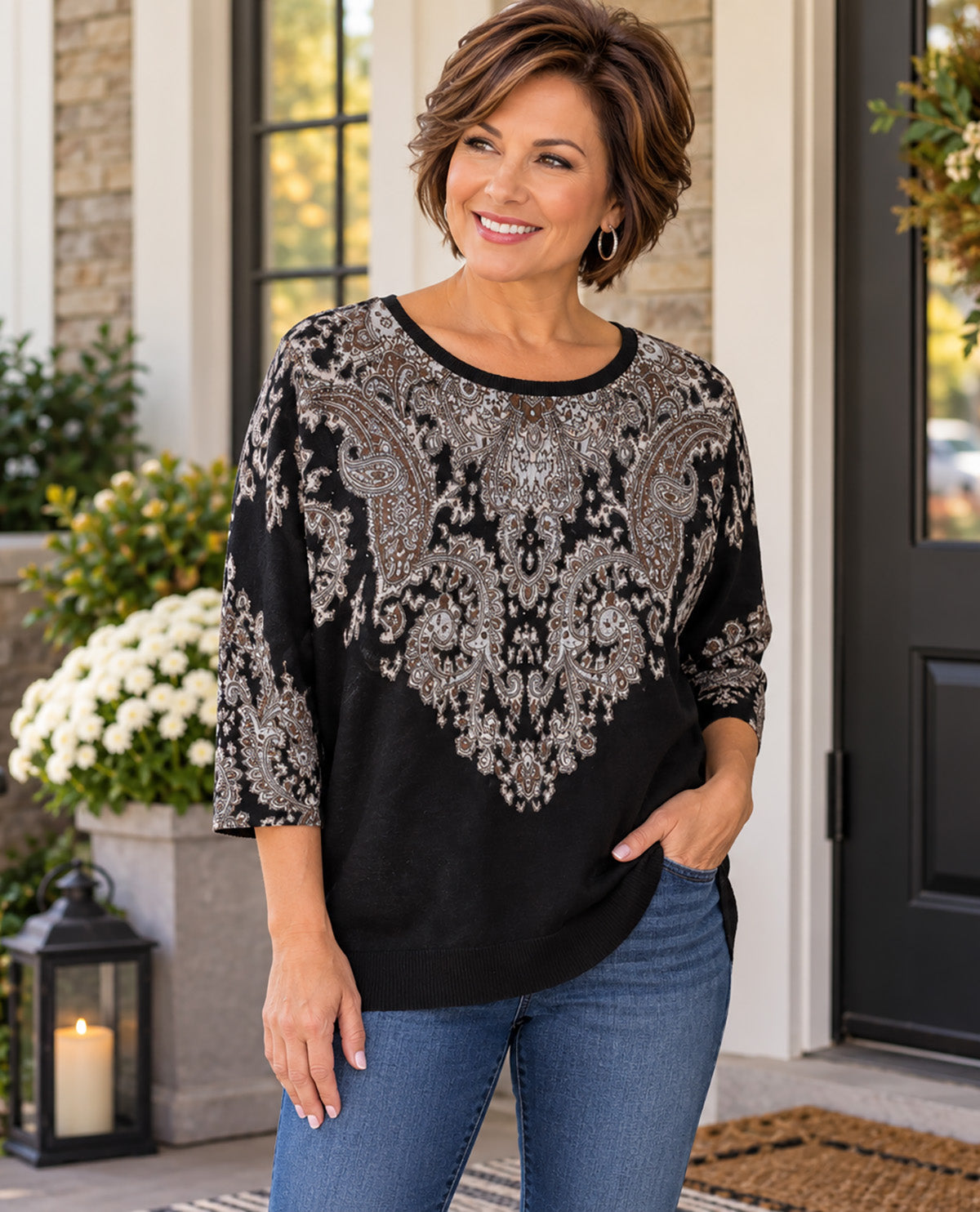 A woman wearing a black three-quarter sleeve top with a large silver paisley pattern on the chest and sleeves, paired with blue jeans, standing on a front porch.