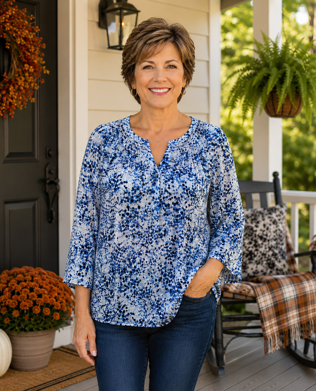 A smiling woman stands on a porch wearing a blue and white floral print long-sleeve blouse with dark blue jeans. The porch has a dark door, a hanging wreath, a rocking chair with a plaid throw, and greenery in the background.