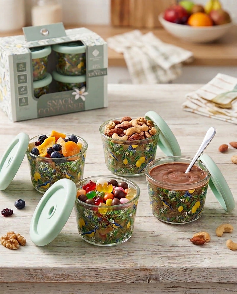 Four clear glass snack containers with pale mint green lids are arranged on a rustic wooden table, filled with dried fruit, nuts, and a chocolate spread. The product box is visible in the background along with a softly blurred kitchen setting.