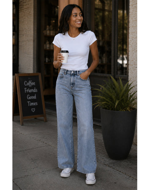 Woman in white t-shirt and blue jeans holding a coffee cup outdoors.