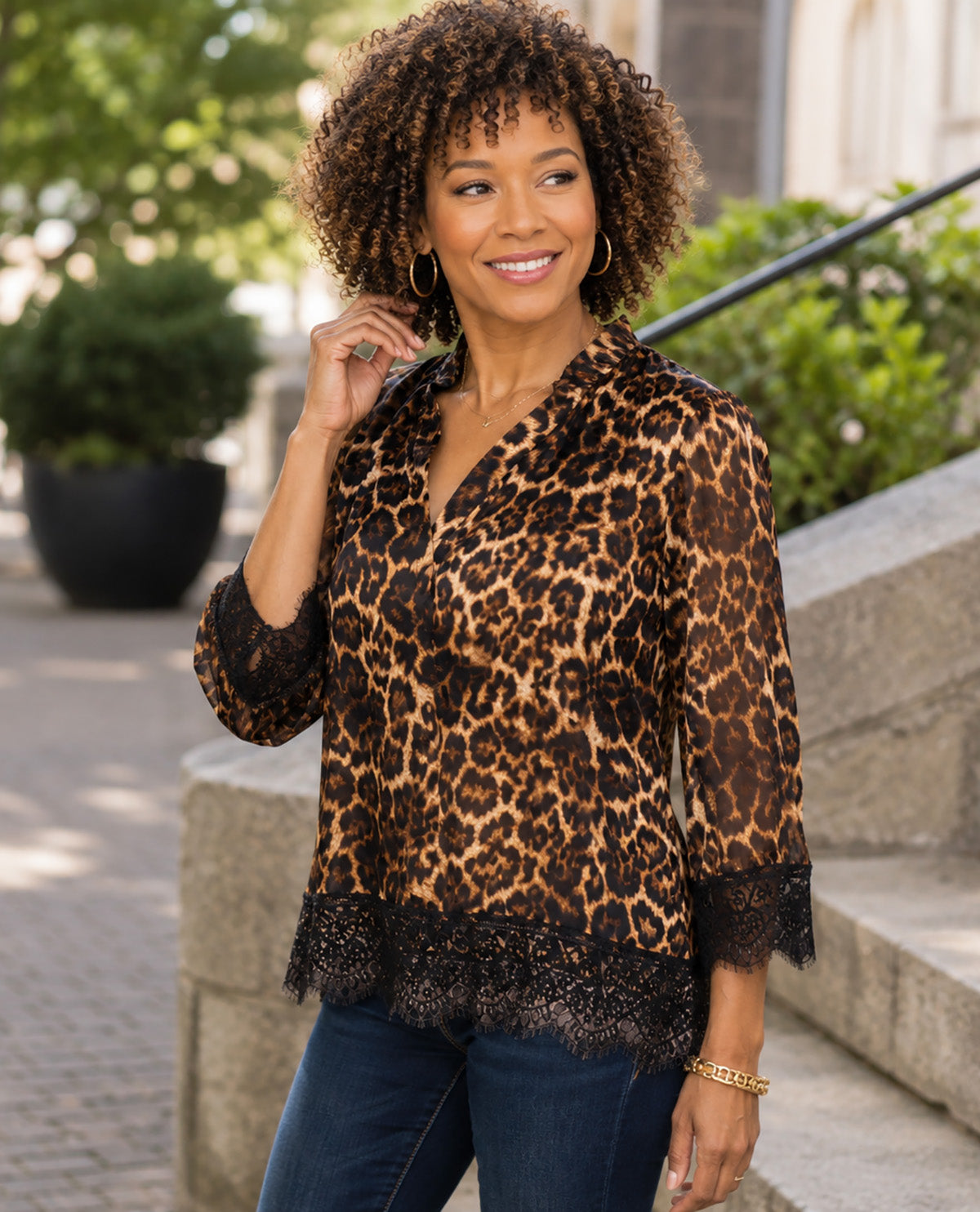 A woman wearing a leopard print blouse with black lace trim on the cuffs and hem, paired with blue jeans, standing outdoors on stone steps.
