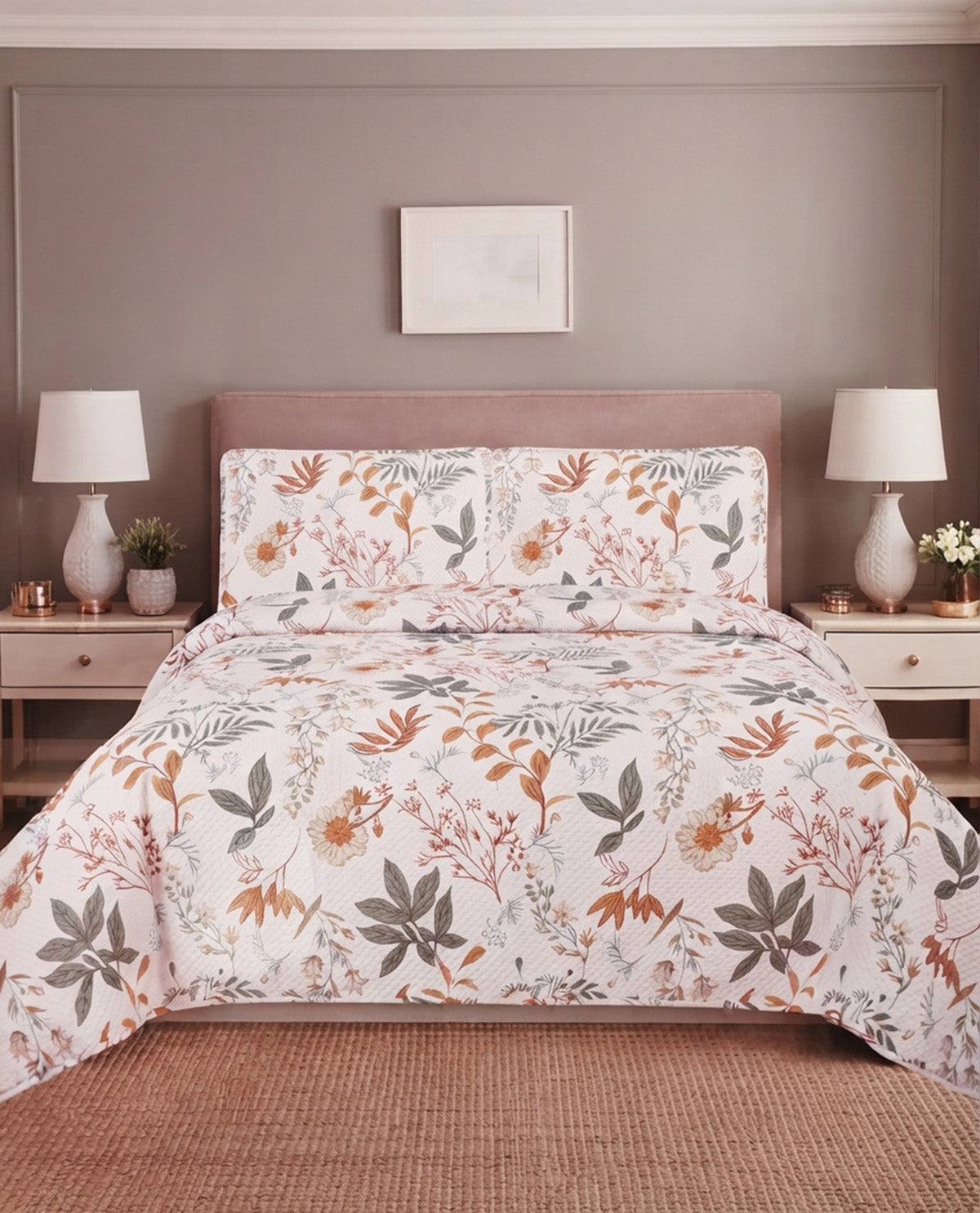 A bed dressed in a white floral comforter and matching pillow shams, styled in a bedroom with a mauve headboard, bedside tables, lamps, and a woven rug.
