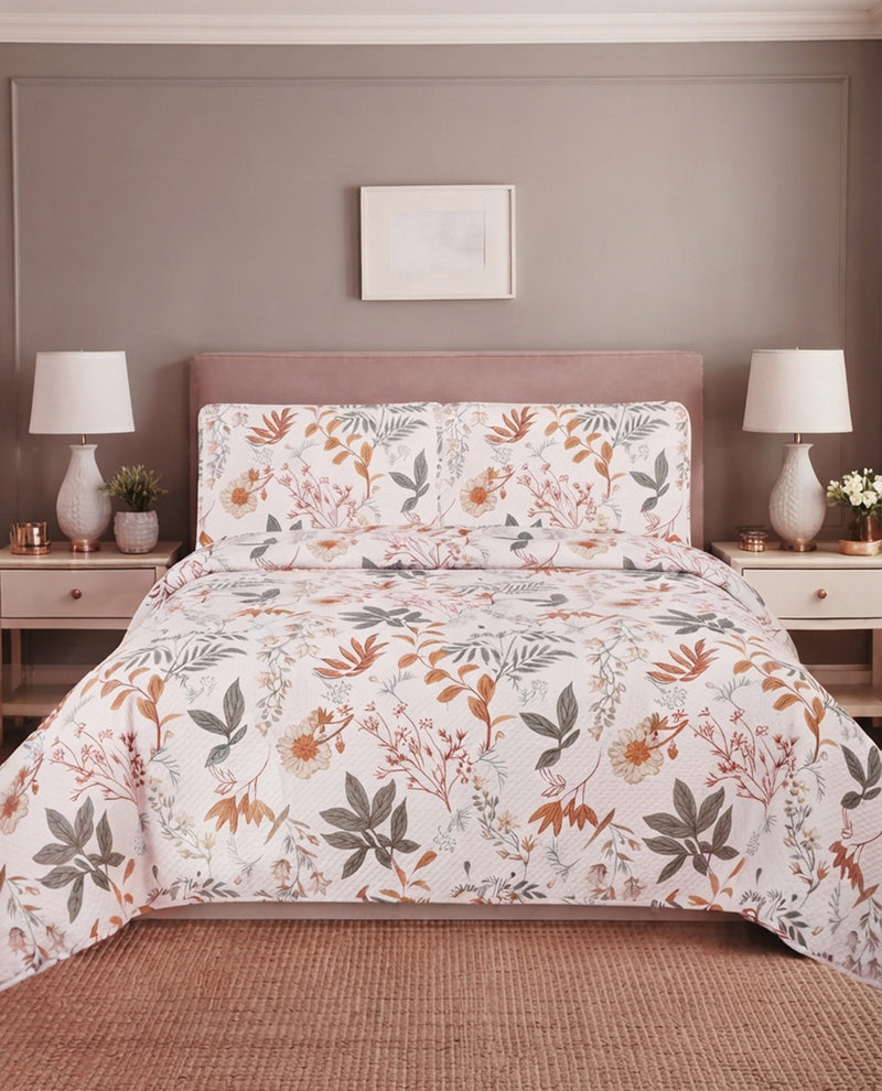 A bed dressed in a white floral comforter and matching pillow shams, styled in a bedroom with a mauve headboard, bedside tables, lamps, and a woven rug.