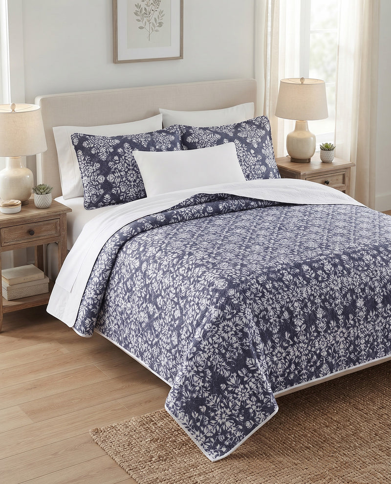 A neatly made bed in a bright bedroom setting, featuring a navy blue and white floral patterned comforter/duvet with matching pillow shams, white accent pillows, and white sheets. The bed is flanked by wooden nightstands with lit table lamps, a light wood floor, and a woven rug.
