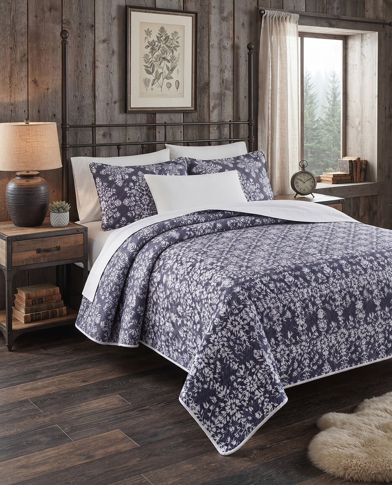 A bed dressed in a navy and white floral-patterned quilt and matching pillow shams, styled in a rustic bedroom with dark wood walls, nightstands, a lamp, and a window.
