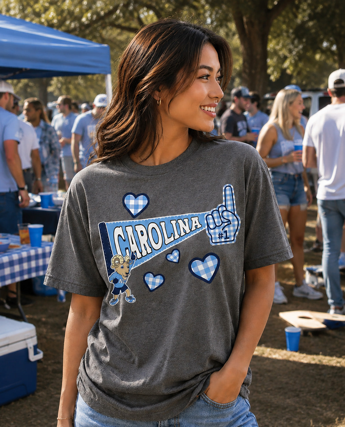 A woman wearing a dark gray Carolina-themed graphic t-shirt with blue and white gingham hearts, a blue hand gesture, and a cartoon character, standing outdoors at a festival or tailgate event.
