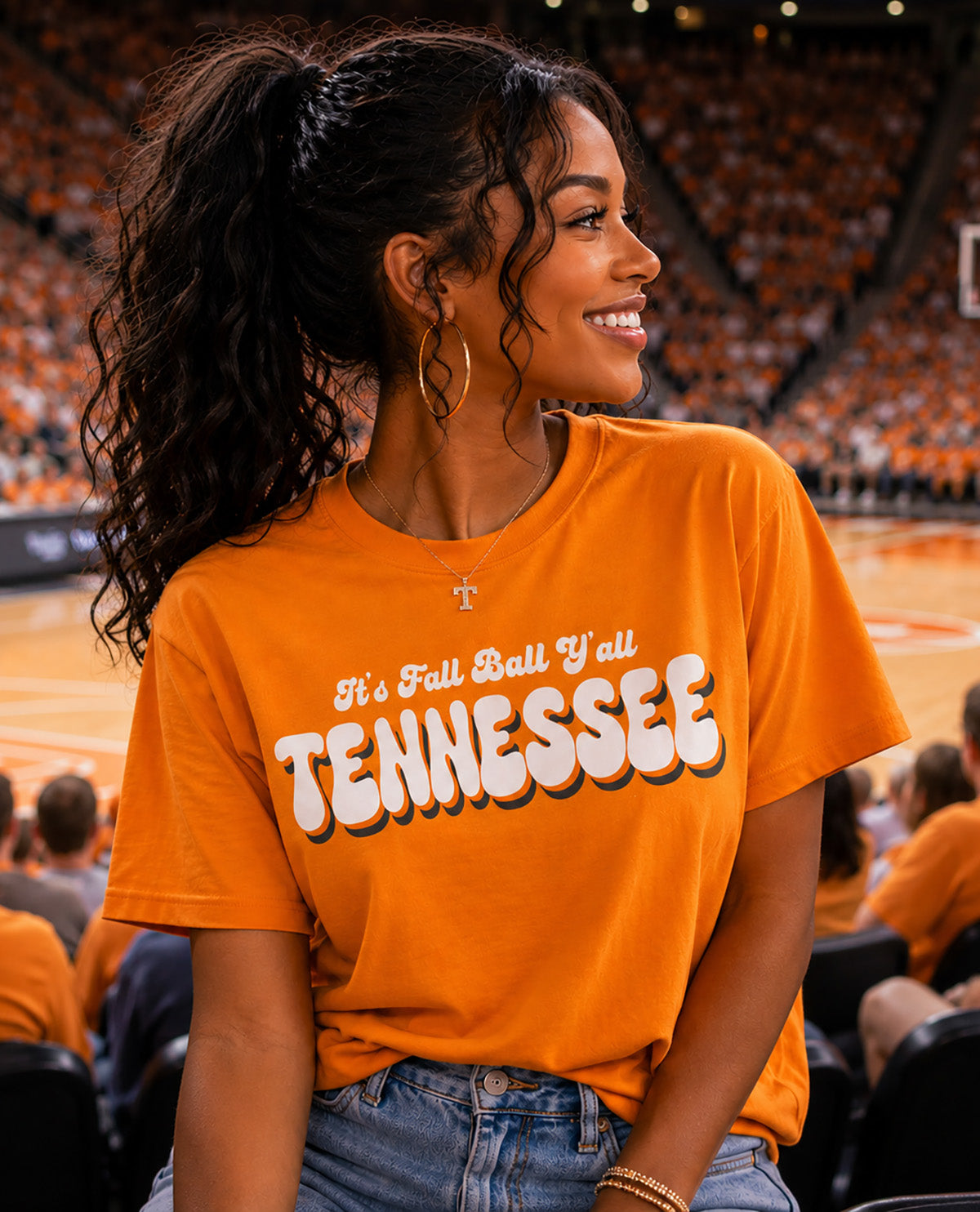 A woman in a bright orange Tennessee basketball jersey sits in a crowded stadium, smiling and looking to the side. The jersey features the text 'It's Fall Ball Y'all TENNESSEE' in white with black outlines.