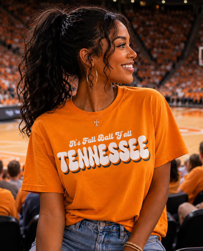 A woman in a bright orange Tennessee basketball jersey sits in a crowded stadium, smiling and looking to the side. The jersey features the text 'It's Fall Ball Y'all TENNESSEE' in white with black outlines.