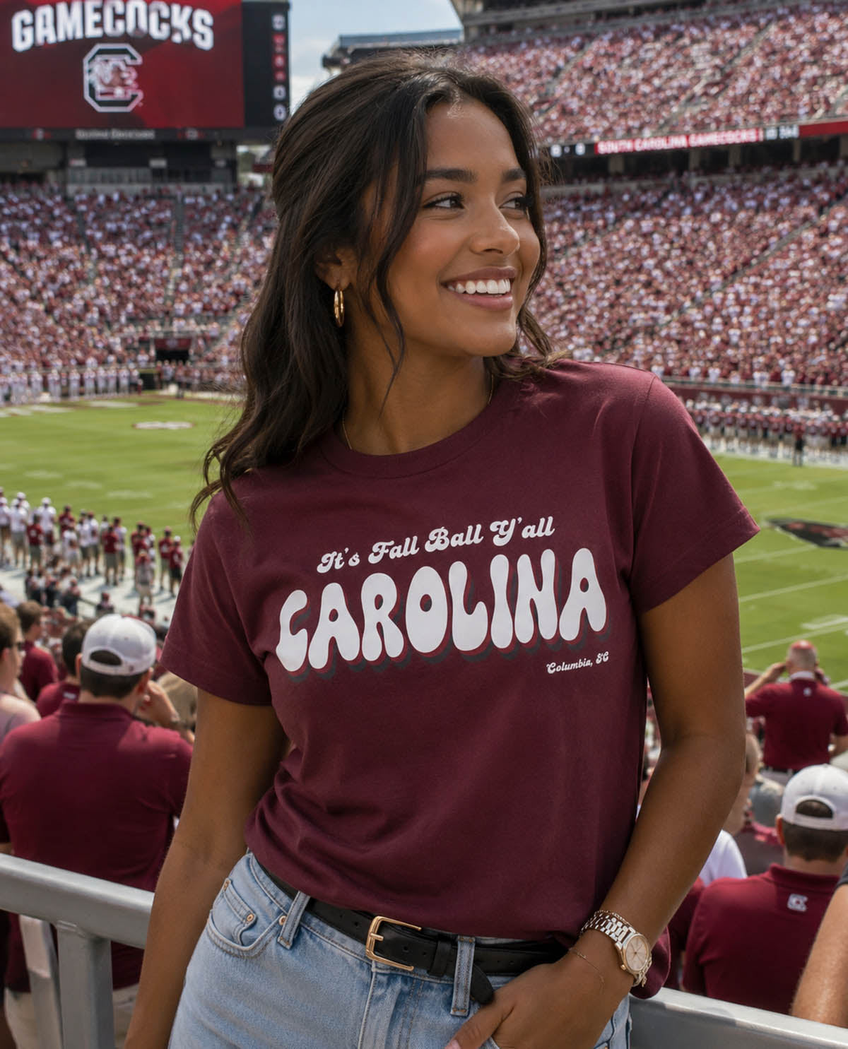 Woman wearing a maroon 'CAROLINA' t-shirt at a sports event with a crowd in the background.