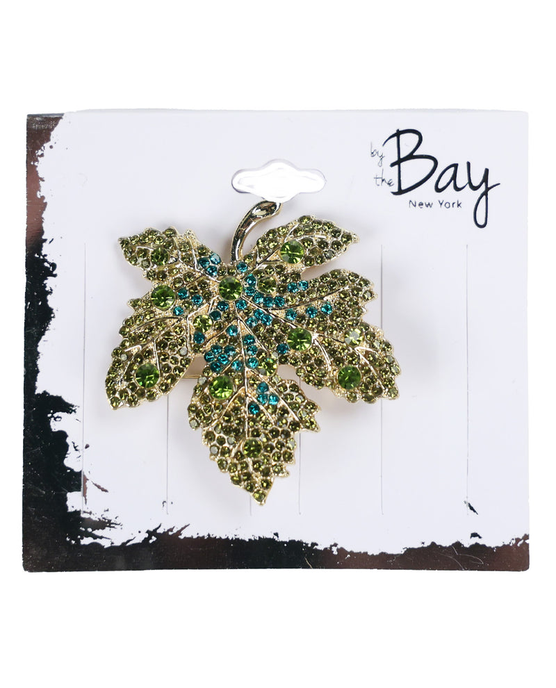 A gold-toned leaf-shaped brooch embellished with green and blue rhinestones, displayed on a white card with a hanger slot. The brand name 'by the Bay New York' is printed in black script on the top right of the card.