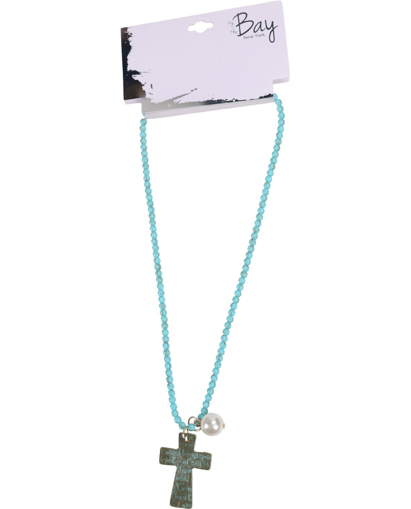 A turquoise blue beaded necklace with a textured dark green cross pendant and a small white pearl, displayed on a white card labeled 'Bay' against a white background.