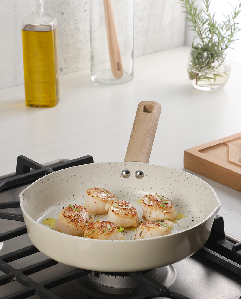 Scallops being cooked in a beige frying pan on a stovetop with a wooden spatula and oil bottle in the background.