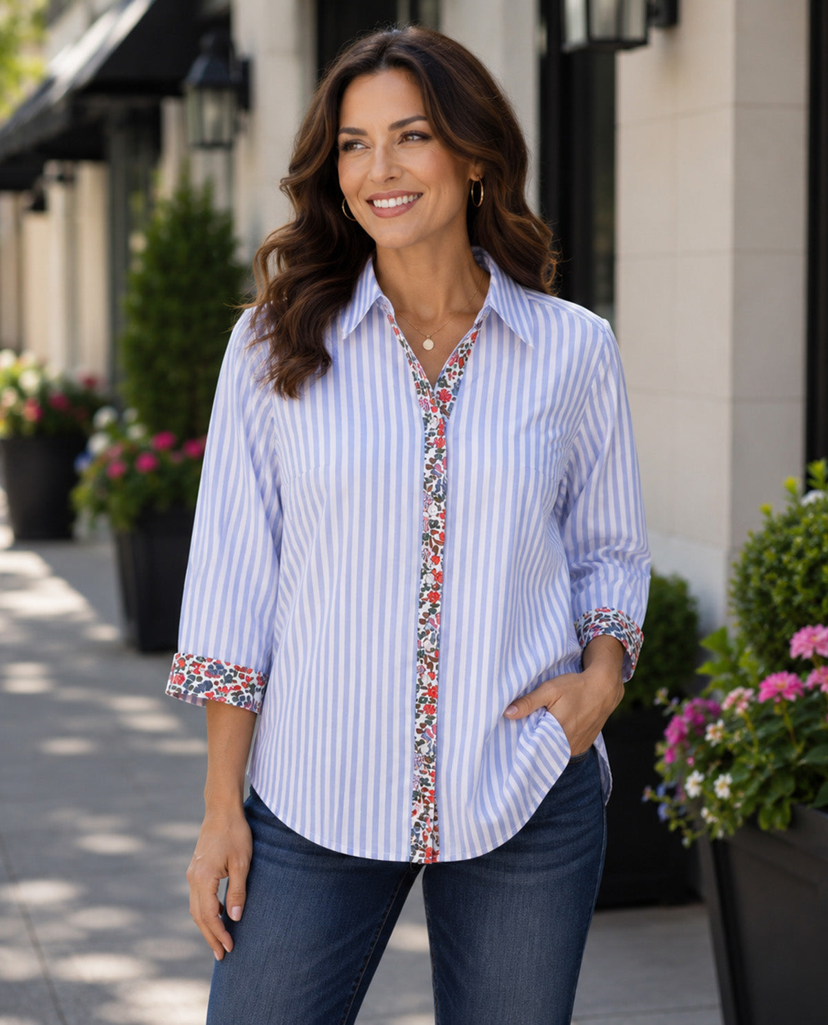 A woman wearing a light blue and white vertically striped button-down shirt with a colorful floral trim down the front placket and on the rolled-up sleeves, paired with dark blue jeans, standing outdoors on a sidewalk.