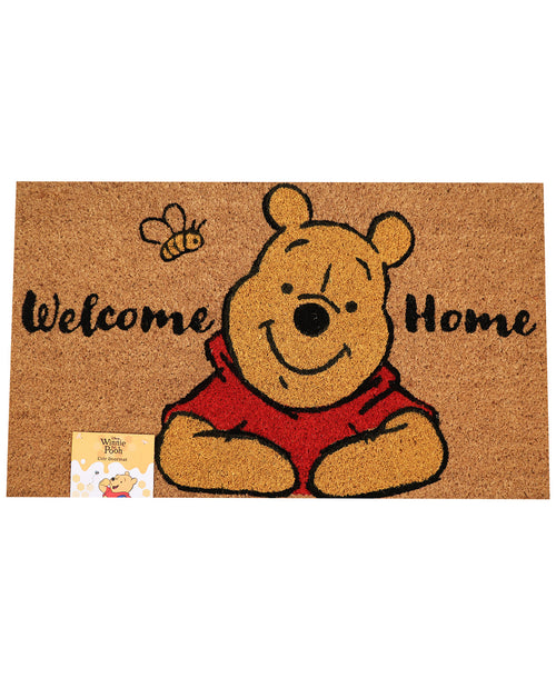 A rectangular coir doormat featuring Winnie the Pooh, with the words 'Welcome Home' in black script and a small bee illustration. A small Winnie the Pooh card is attached to the lower left corner.