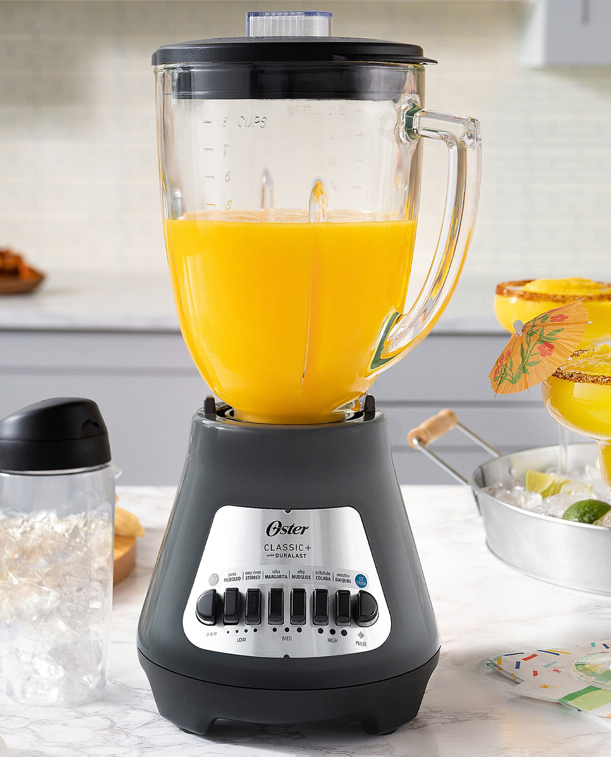 An Oster Classic 4 in 1 blender filled with orange juice, set on a marble countertop in a kitchen environment with cocktail glasses and a shaker bottle in the background.