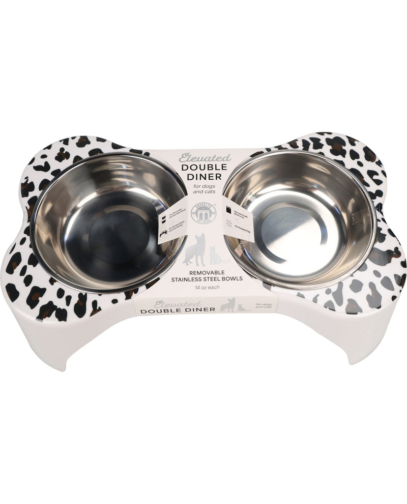 A pet double-diner bowl set featuring two stainless steel bowls set into a white elevated base with a black and white cow-print pattern, shown on a white background.