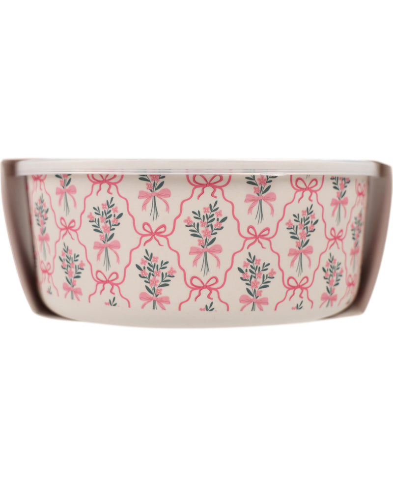 A round ceramic bowl with a pink and green floral pattern featuring bows and ribbon motifs on a white background, shown inside its brown cardboard packaging sleeve.