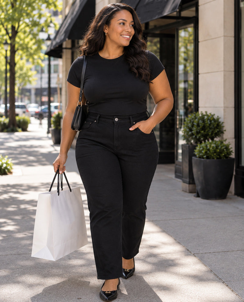 A woman walking on a city sidewalk wearing a black short-sleeve crewneck t-shirt and black high-waisted straight-leg jeans, holding a white paper shopping bag.