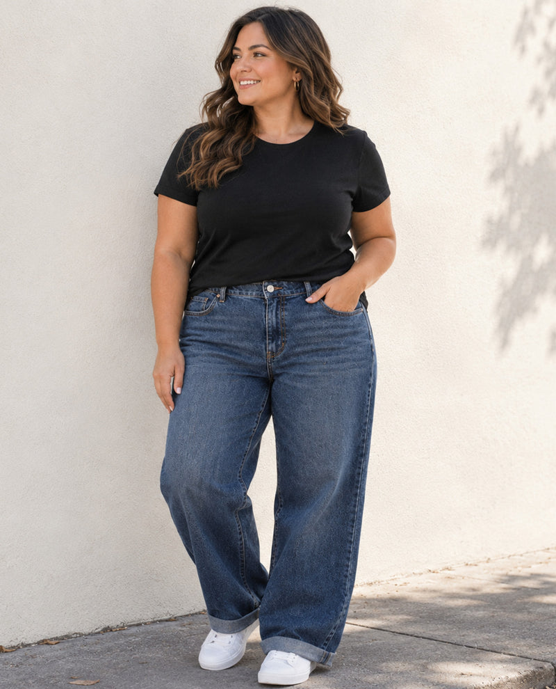 A woman wearing a black short-sleeve t-shirt, wide-leg blue jeans, and white sneakers, standing against a light beige stucco wall.