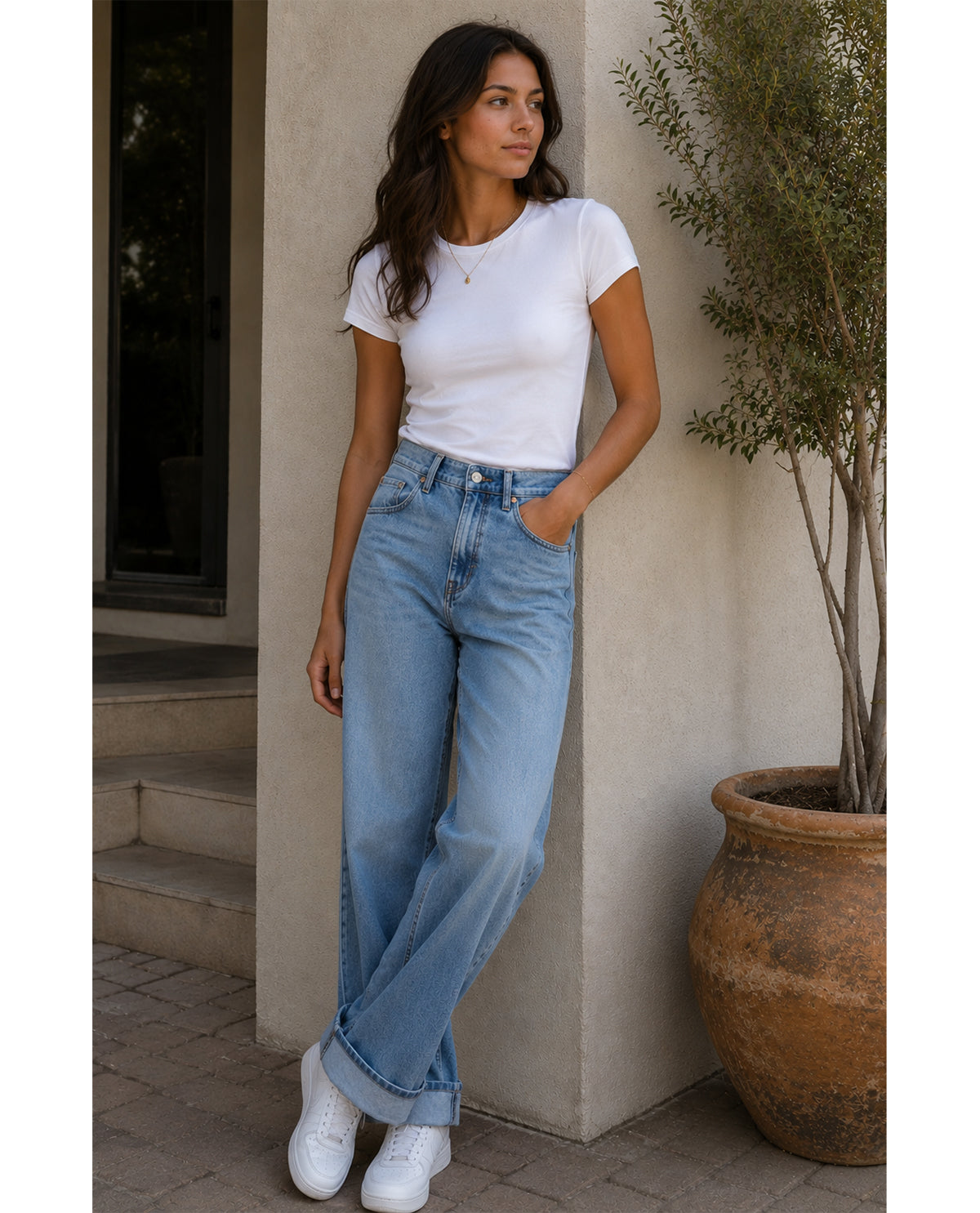 Woman wearing a white t-shirt and blue jeans standing against a beige wall.