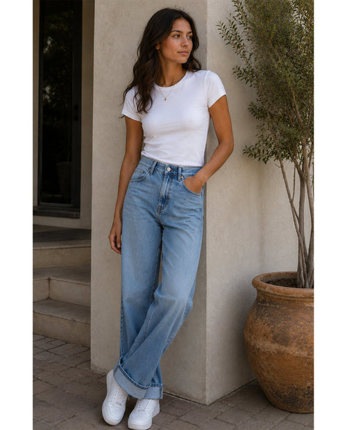 Woman wearing a white t-shirt and blue jeans standing against a beige wall.