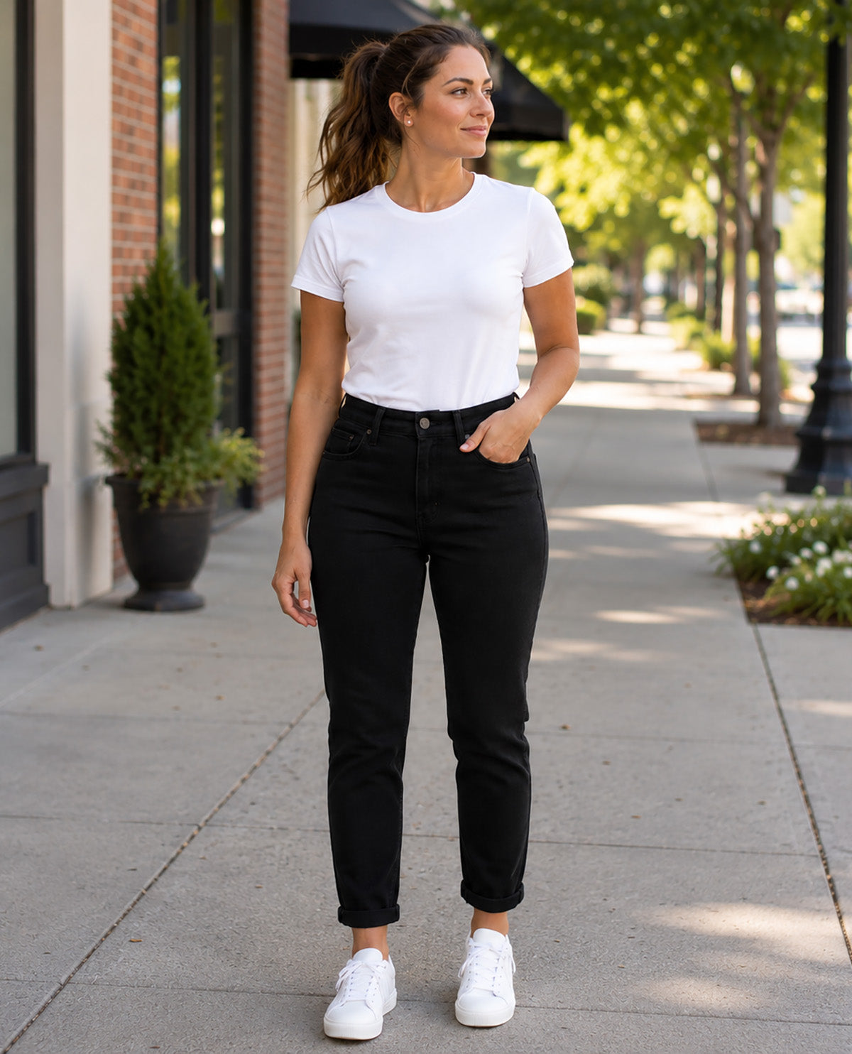 A woman stands on a city sidewalk wearing a white short-sleeve crewneck t-shirt, black high-waisted ankle-length jeans, and white sneakers.