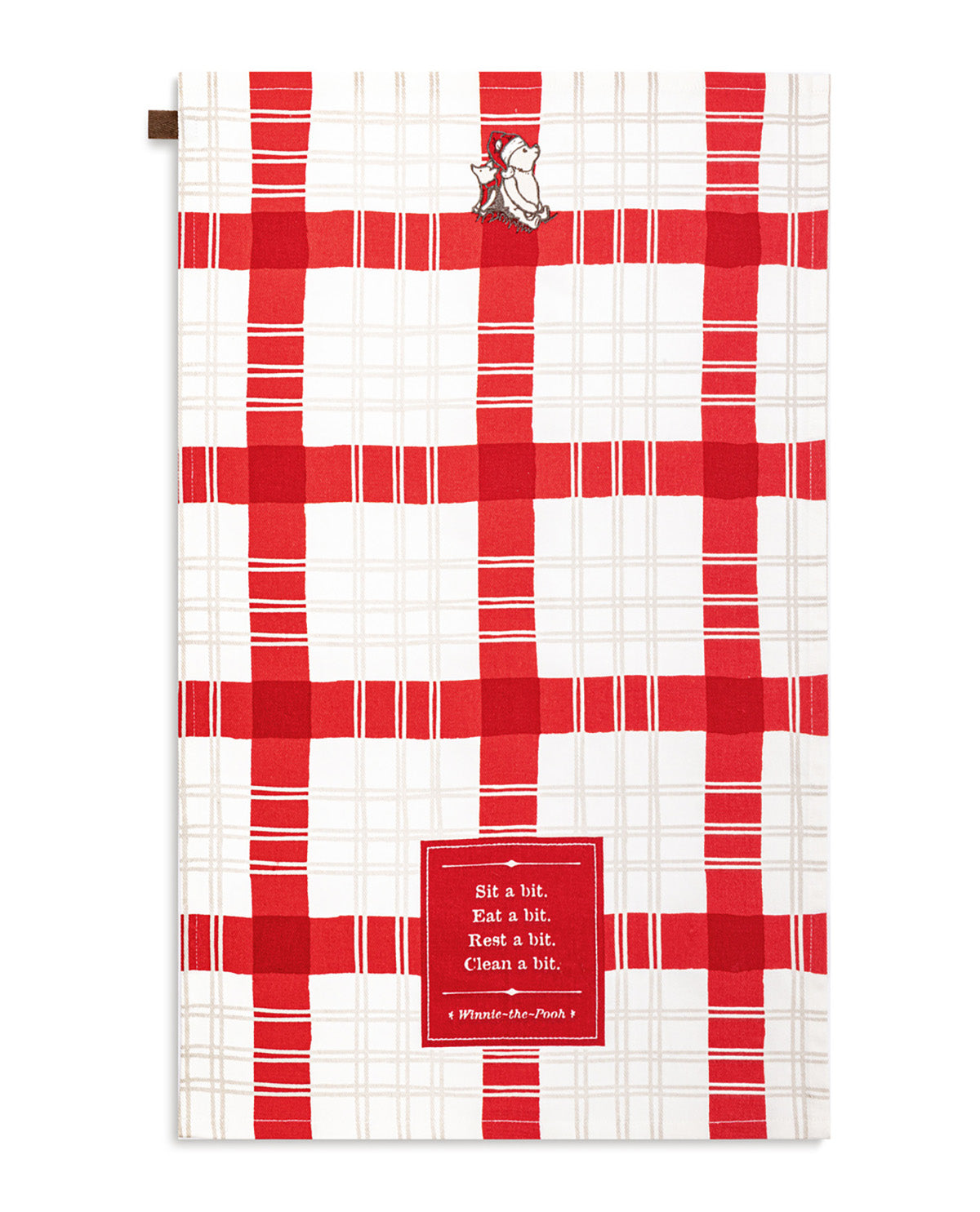 A kitchen towel featuring a red and white plaid pattern, with a small Winnie-the-Pooh character near the top and a red square label at the bottom that reads, 'Sit a bit. Eat a bit.
