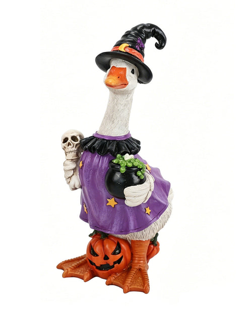 A Halloween decorative figurine of a goose dressed as a witch, wearing a black hat and purple dress, holding a skull and a cauldron, standing on a carved pumpkin against a white background.