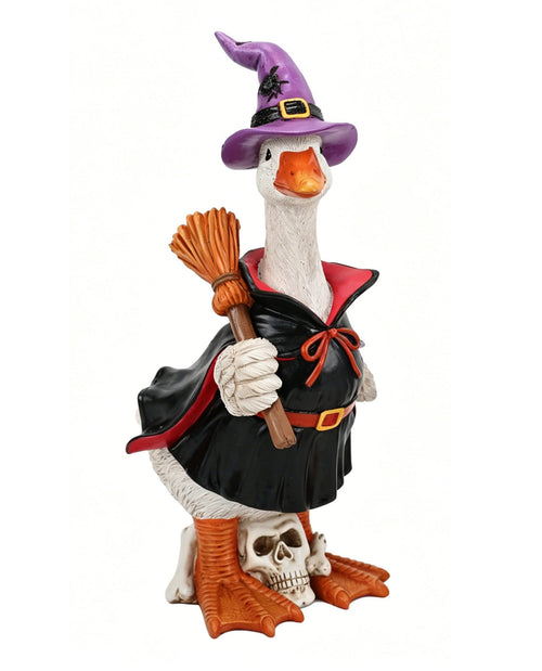 A whimsical Halloween figurine of a goose dressed as a witch, wearing a purple hat, black cape with red trim, orange boots, and holding a broomstick, standing on a skull against a white background.