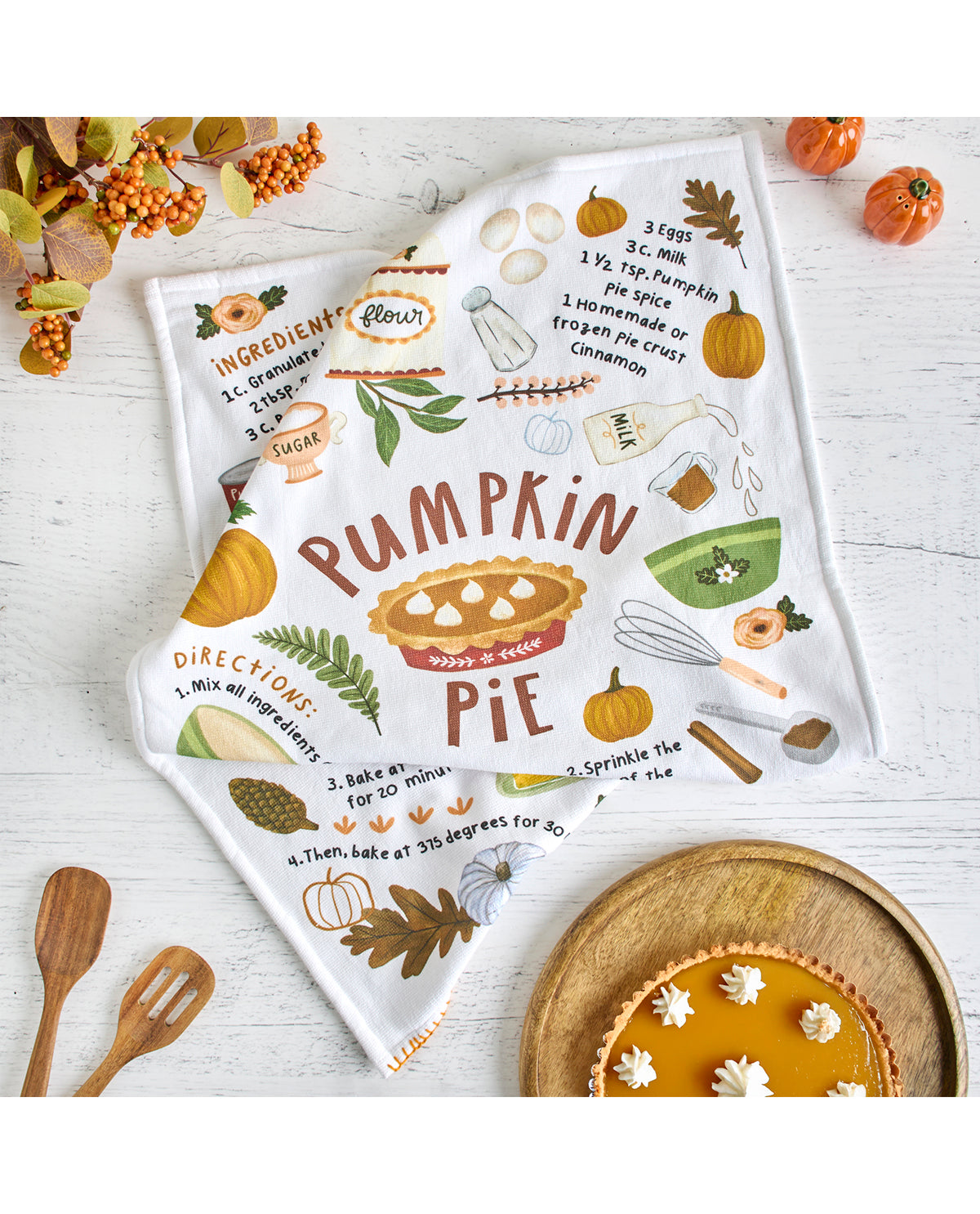 Tea towel with pumpkin pie design and recipe on a white surface with pumpkins and leaves.