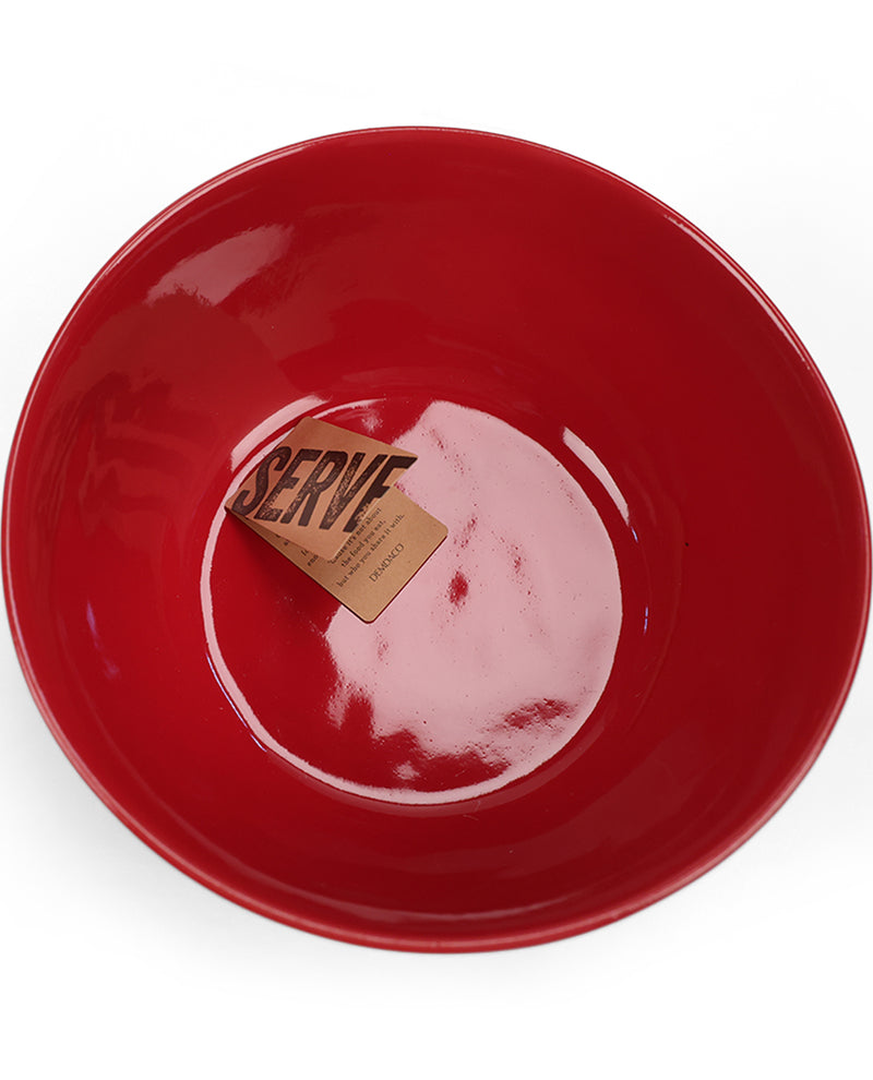 A glossy red ceramic bowl photographed from above on a white background, with a small paper tag inside the bowl that reads “SERVE.”.