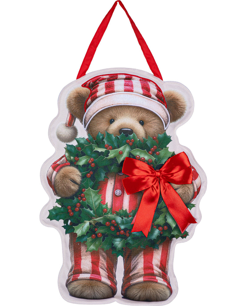 A Christmas hanging ornament shaped like a teddy bear wearing red and white striped pajamas and a Santa hat, holding a green holly wreath with red berries and a large red bow. A red hanging loop is attached at the top.