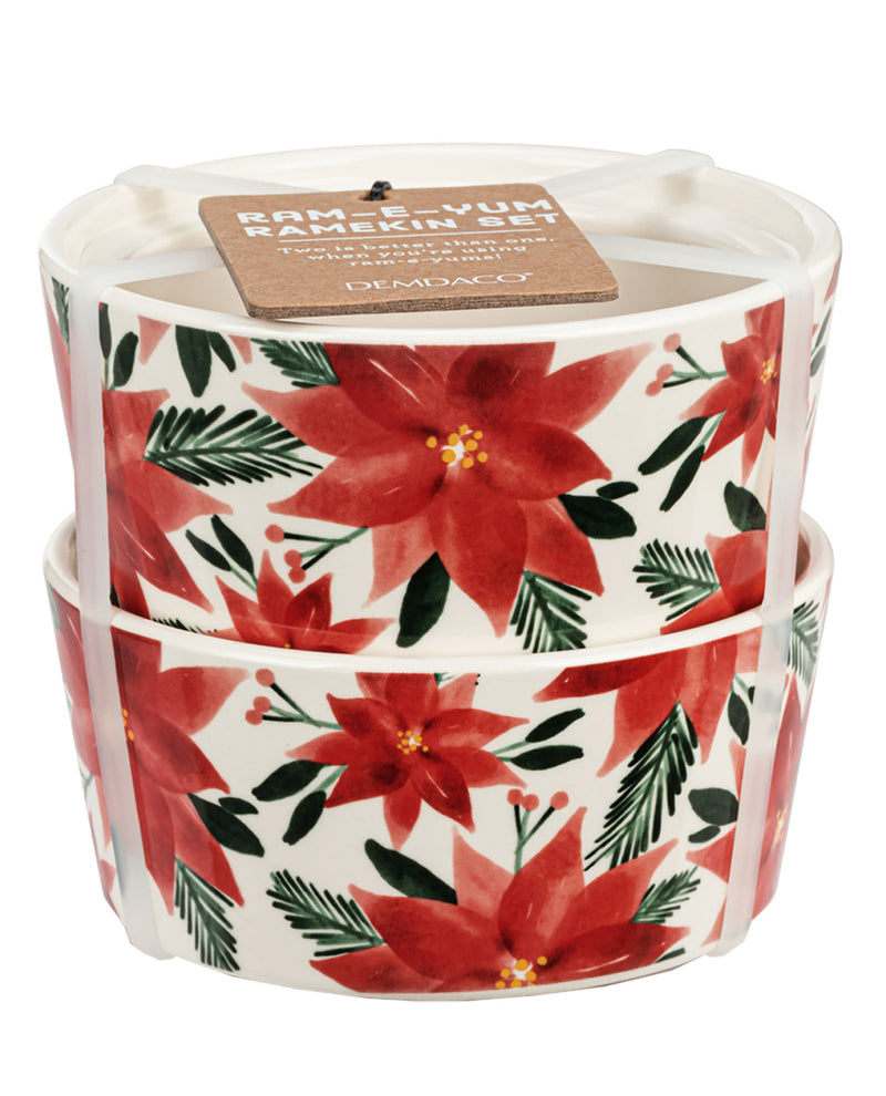 A set of two stacked ramekins with a white base decorated with a red poinsettia and green pine branch pattern. A brown paper tag is attached to the top candle lid.