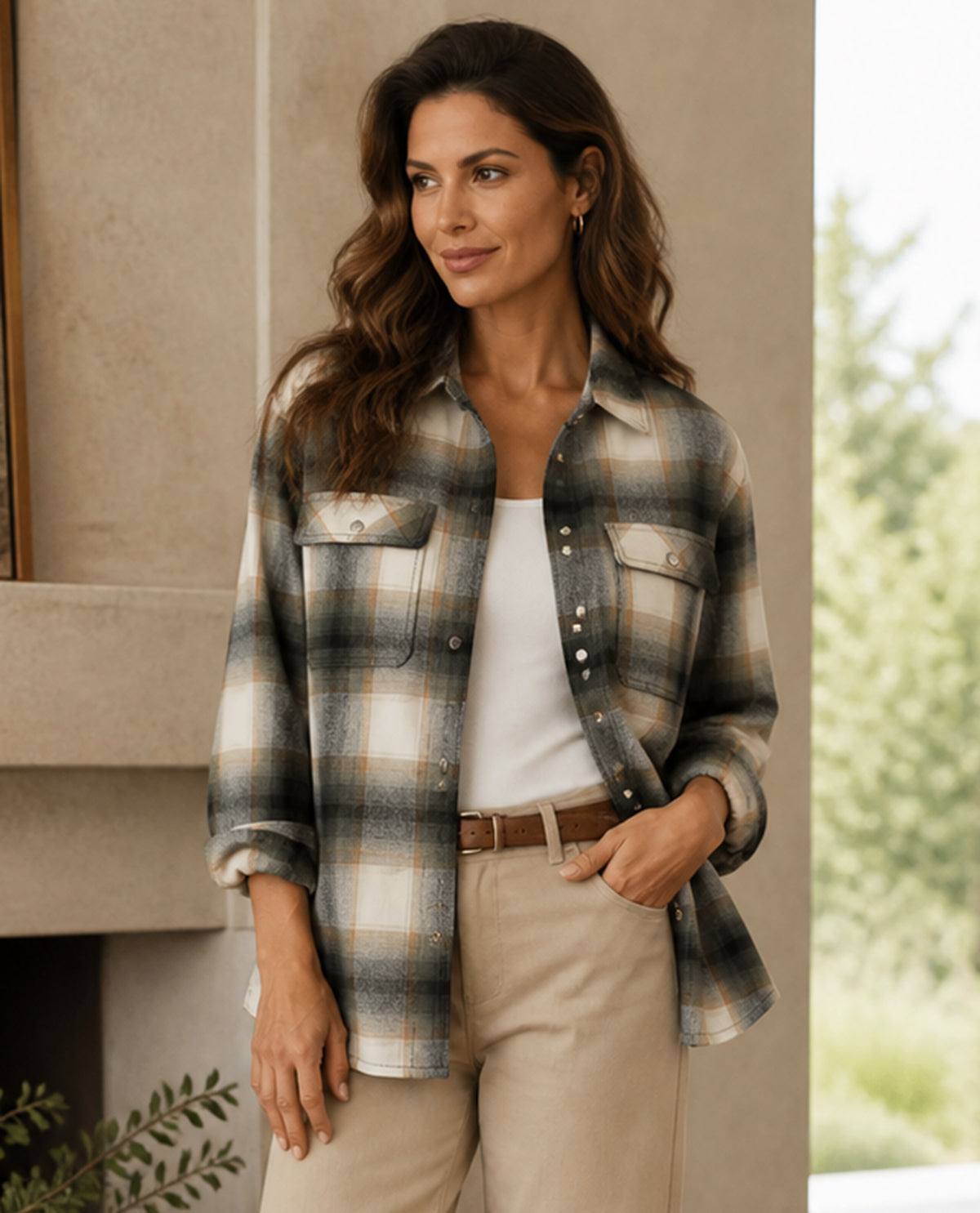 Woman wearing a plaid shirt over a white top and beige pants, standing indoors with a blurred outdoor background.