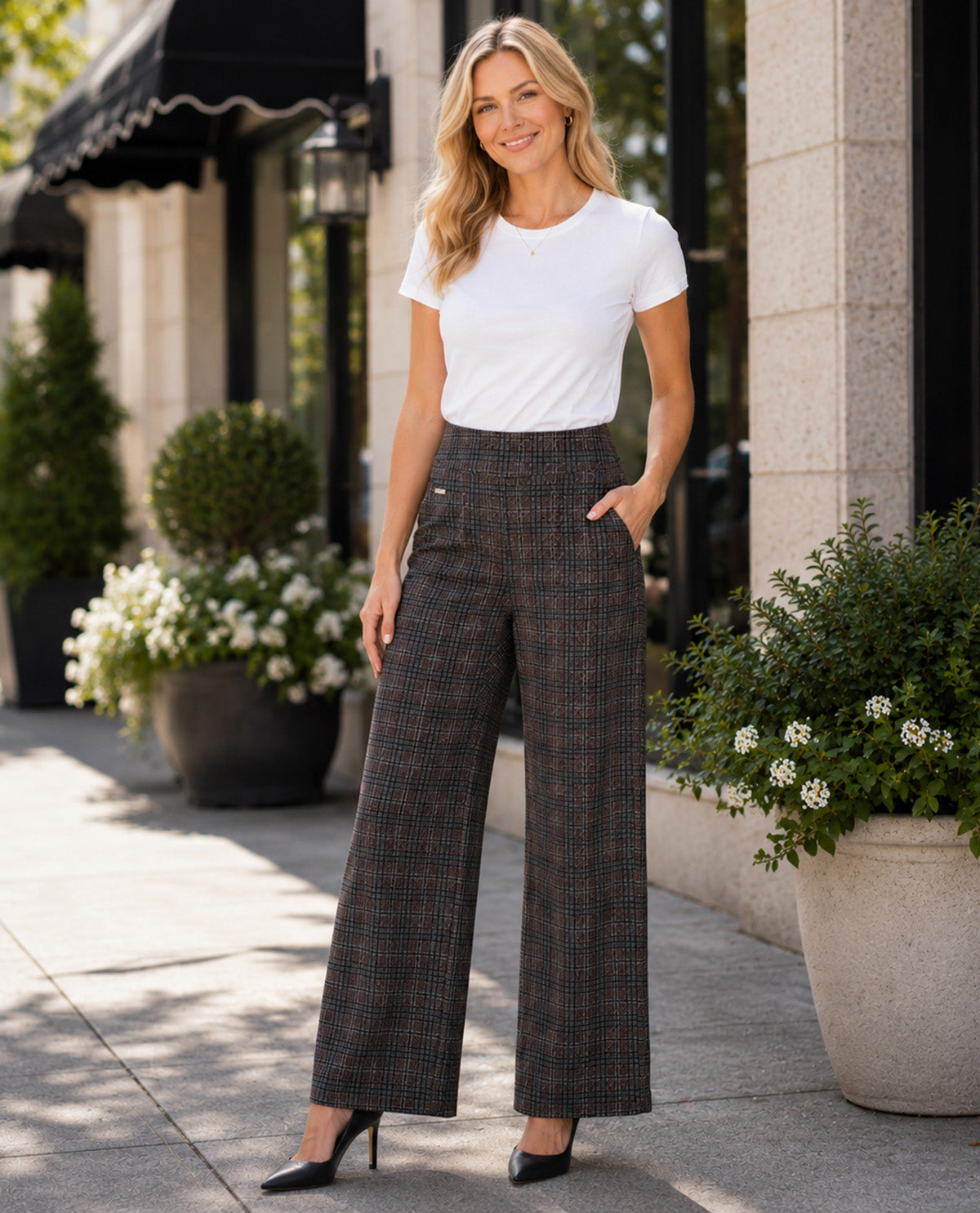 A woman stands on a city sidewalk wearing a white short-sleeve crewneck T-shirt and high-waisted, wide-leg plaid trousers in dark gray and brown tones, paired with black heels.