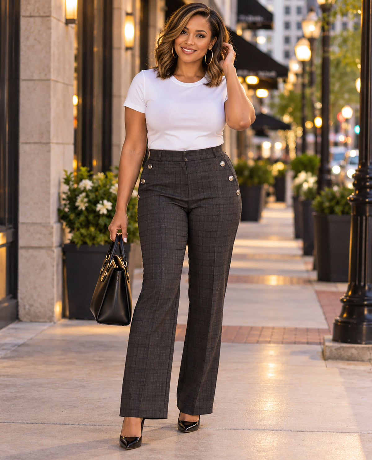 A woman stands on a city sidewalk wearing a white short-sleeve crewneck t-shirt and high-waisted dark gray plaid trousers with decorative gold buttons on the hips. She is holding a black handbag and wearing black pointed-toe heels.