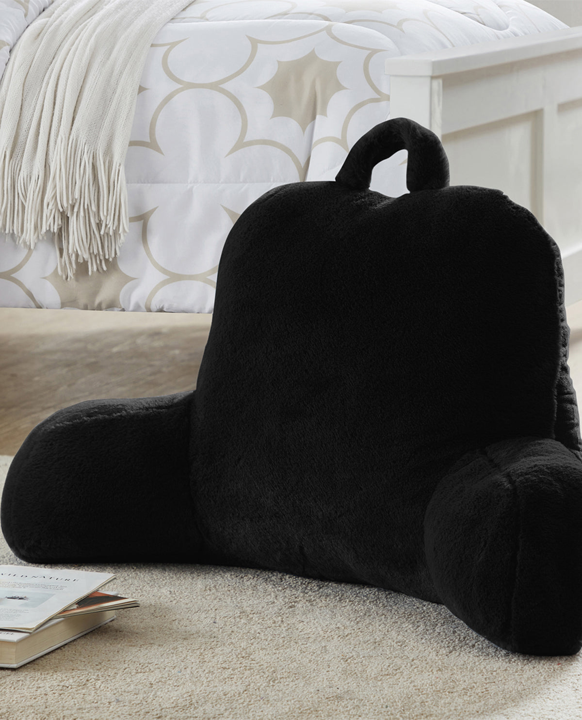 Black pillow with armrests on a carpeted floor next to a book, with a patterned wall in the background.