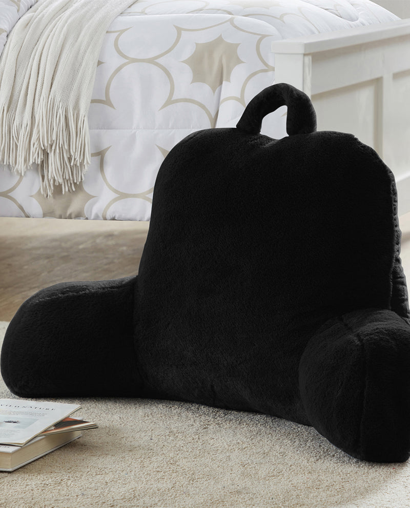 Black pillow with armrests on a carpeted floor next to a book, with a patterned wall in the background.
