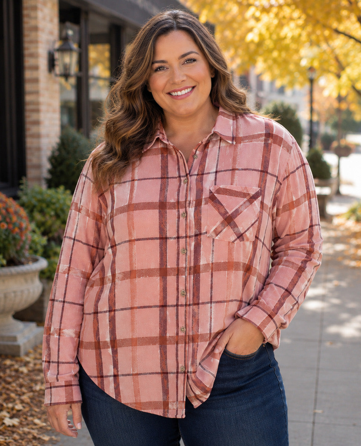 A woman wearing a pink and rust plaid button-down shirt with dark blue jeans, standing outdoors on a sidewalk with autumn trees in the background.