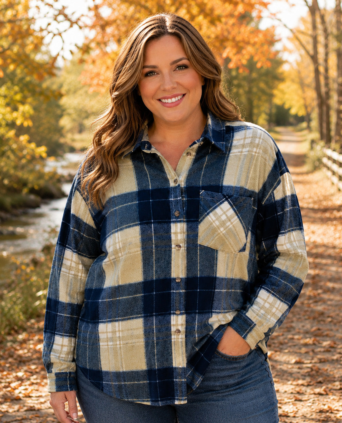 A woman smiling outdoors in a long-sleeve plaid button-up shirt with blue and beige tones, paired with blue jeans, set against a blurred autumn path with trees and a stream.