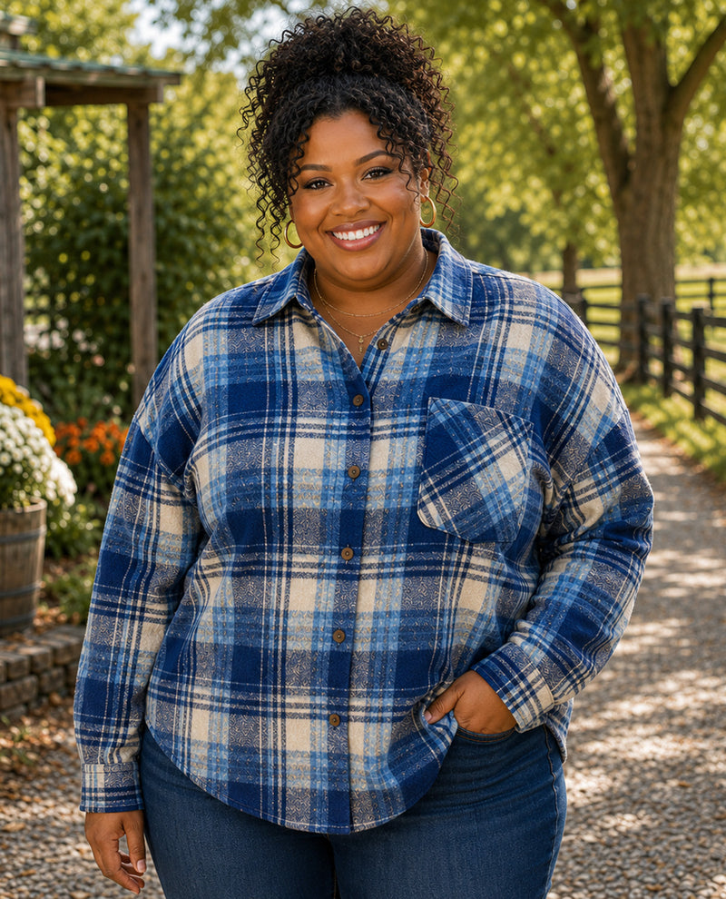 A smiling woman with curly hair stands outdoors on a gravel path wearing a blue and cream plaid button-up flannel shirt and dark blue jeans.